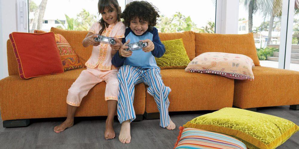 two latino kids playing a game while sitting on an orange couch in a room with carpet flooring